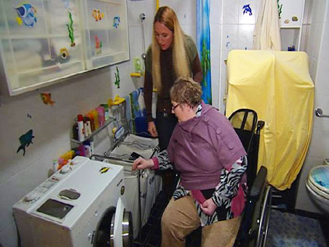 Klientin im Rollstuhl und Betreuerin füllen Schmutzwäsche in die Waschmaschine
