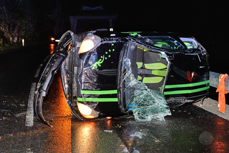 Schwarzgrüner Kleinwagen liegt mit kaputter Windschutzscheibe umgestürzt auf regennasser Straße