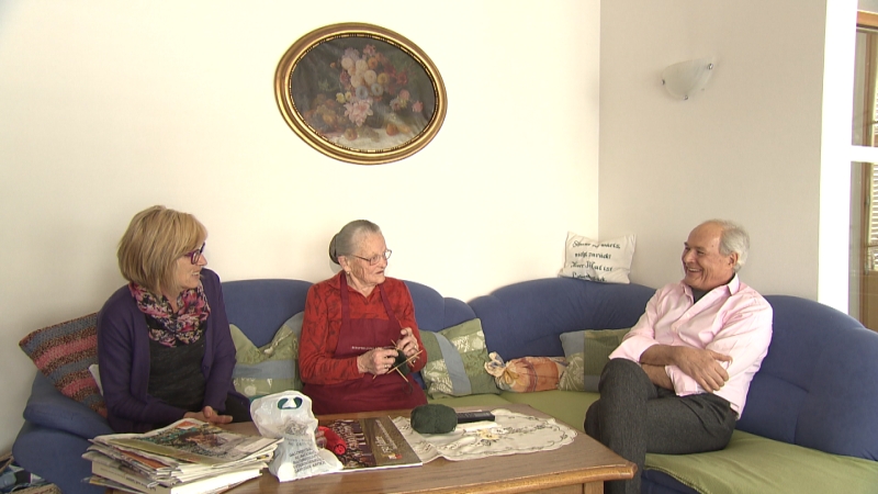 Reinhard Zingerle im Gespräch mit zwei Frauen von Fam. Pfeifer auf Sofa, eine Frau strickt Socken