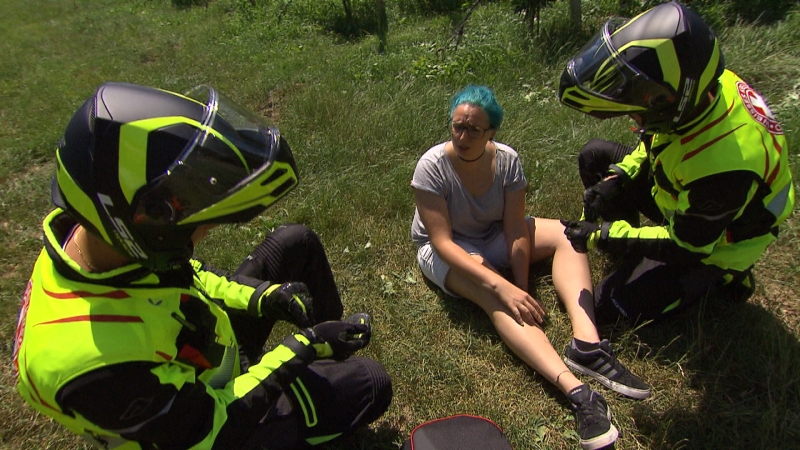 Zwei Motorradsanitäter sprechen mit einer Patientin, die am Boden einer Wiese sitzt (Szene gestellt)