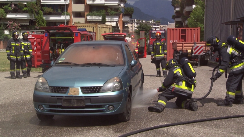 Feuerwehrmänner in Atemschutzmasken lenken auf Kleinwagenmit Schlauch Wasser, aus dem Auto kommt Rauch